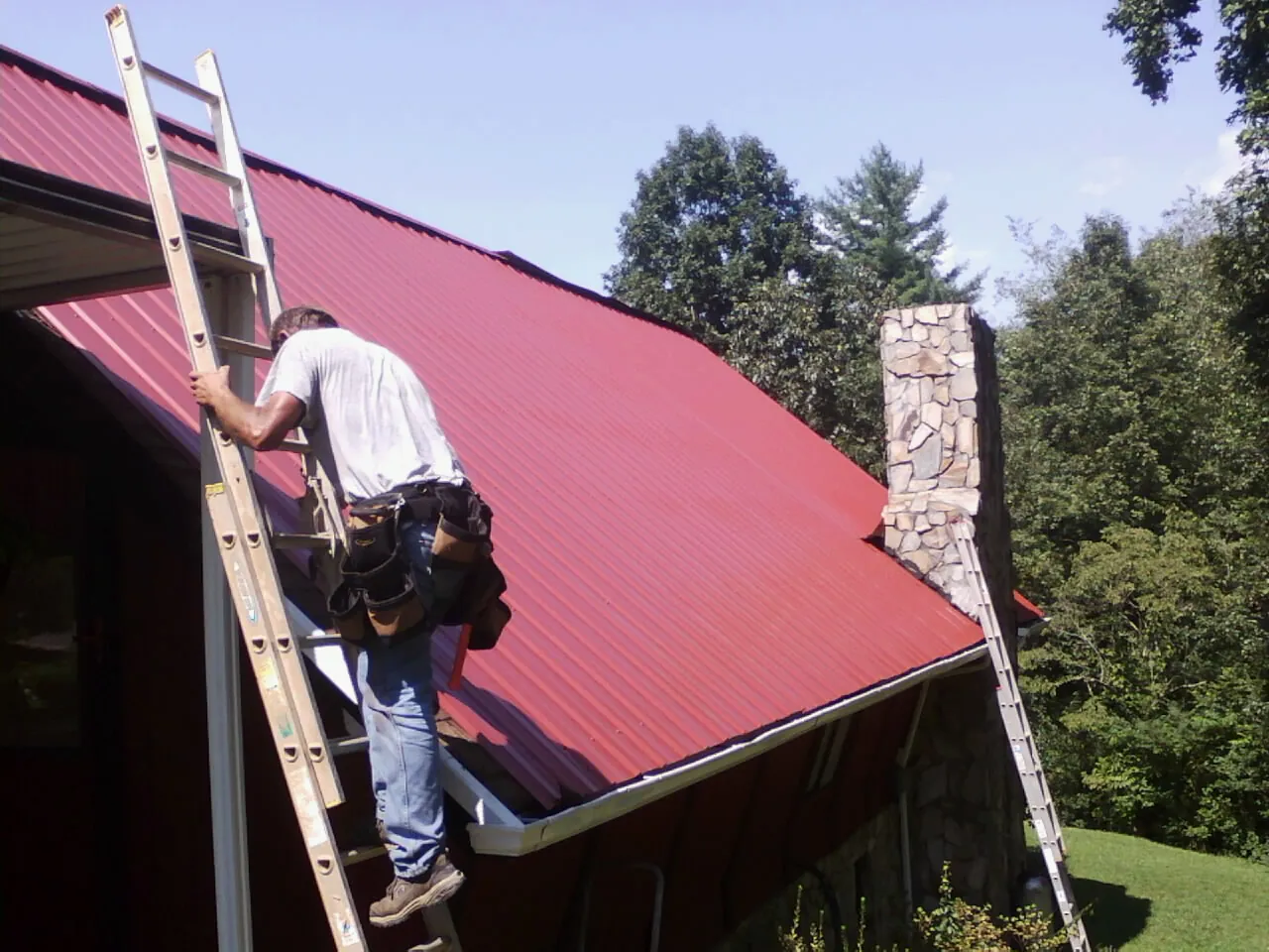 Roofer on a ladder beside a red metal roof and stone chimney — Soffit & Fascia Repair by Roofing Brothers in Williamston
